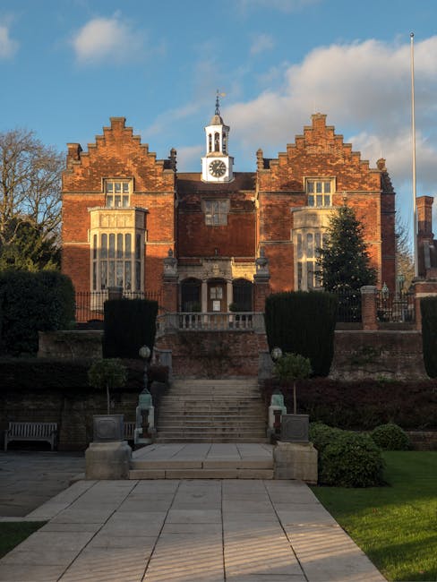 A large historic brick building with Gothic architectural features, including stepped gables, tall narrow windows with stone mullions, and a prominent clock tower with a pointed spire topped by a weat