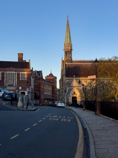 A large historic brick building with Gothic architectural features, including stepped gables, tall narrow windows with stone mullions, and a prominent clock tower with a pointed spire topped by a weat