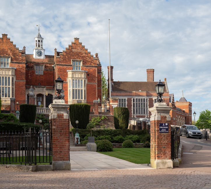 The image displays a stately red brick building with a prominent clock tower and decorative architectural features, situated behind a low brick wall and iron gate. The entrance pathway is paved with s