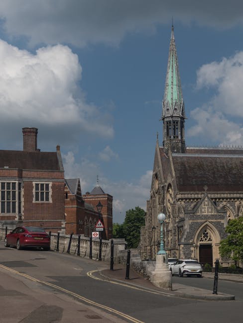 Photograph capturing a street scene near a historic stone church with a tall, green copper spire, situated next to a red-brick building with chimneys. Several vehicles, including a red car parked on t