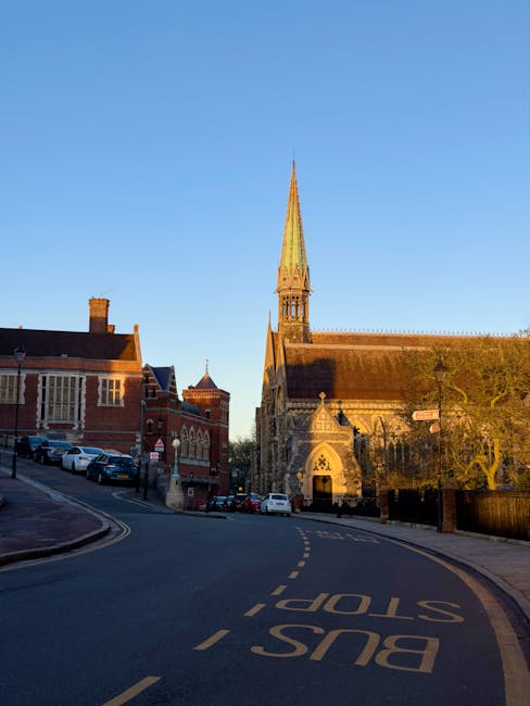 A narrow street in Harrow bordered by historic brick buildings and residential houses, with a large stone church featuring a tall, pointed spire dominating the scene. The street is marked with a 'BUS